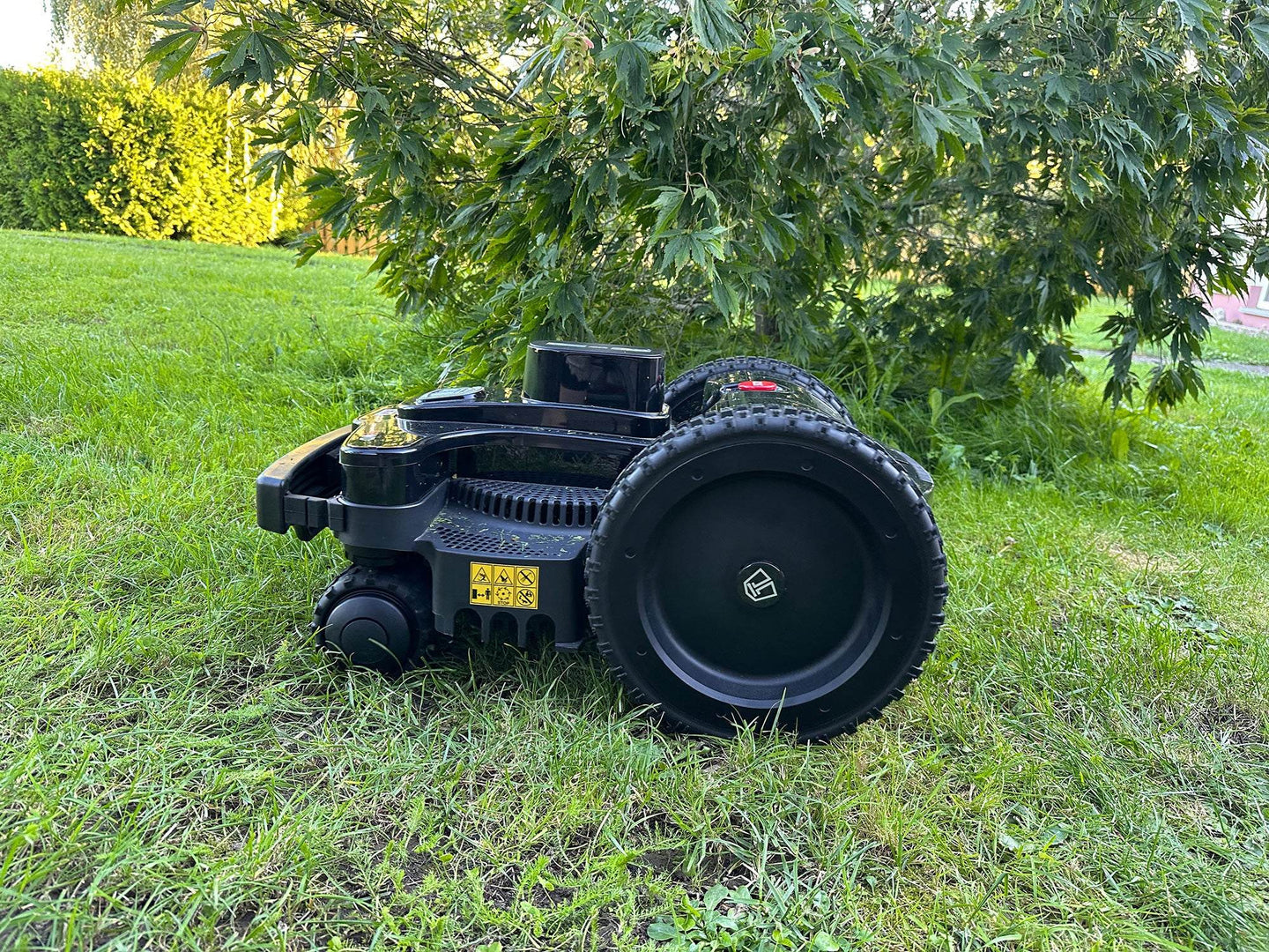 Nahaufnahme des schwarzen Mähroboters von der Seite auf einer Rasenfläche. Das Bild zeigt deutlich die robuste Konstruktion mit dem großen, profilierten Antriebsrad. Auf dem Gehäuse sind gelbe Warnsymbole zu erkennen. Im Hintergrund sieht man unscharfe grüne Vegetation - Blätter eines Baumes oder Strauchs und eine Hecke im Sonnenlicht. Die glänzende schwarze Oberfläche des Roboters reflektiert leicht das Tageslicht.