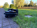 Der schwarze Mähroboter steht auf einem gepflasterten Gartenweg aus grauen, leicht verwitterten Steinplatten. Das Bild zeigt den Roboter von der Seite, wobei besonders die robusten Antriebsräder und das vordere kleinere Stützrad gut zu erkennen sind. Ein Begrenzungsdraht verläuft am Rand des Weges. Im Hintergrund ist saftig grüner Rasen zu sehen, und die Abendsonne taucht den Garten in warmes Licht, was besonders an den Hecken und Büschen im Hintergrund sichtbar ist.