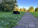 Der schwarze Mähroboter steht im hohen Gras neben einem gepflasterten Gartenweg. Die Aufnahme wurde in der goldenen Abendsonne gemacht, was sich in der warmen Beleuchtung der Bäume und Sträucher im Hintergrund zeigt. Der gepflasterte Weg führt in die Bildtiefe und ist von üppigem Grün gesäumt. Im linken Bildbereich steht ein großer Laubbaum, dessen Blätter im Sonnenlicht leuchten. Der Kontrast zwischen dem dunklen Mähroboter und dem hellen, saftigen Gras ist besonders markant.