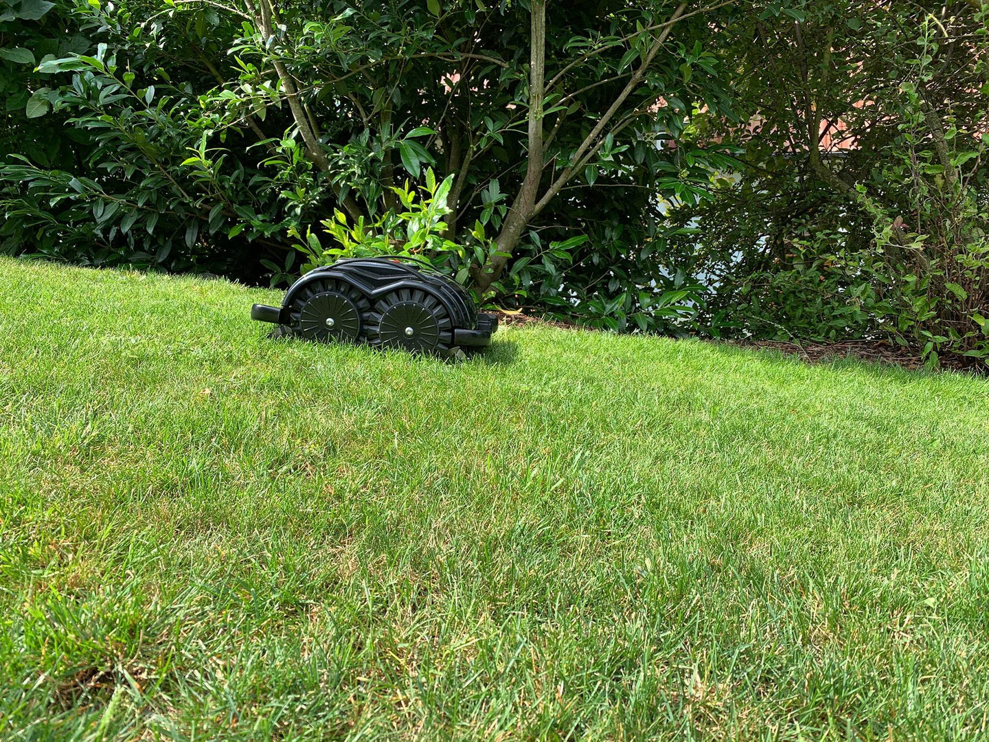 Das Bild zeigt den schwarzen Mähroboter Techline S6, der einen kleinen Abhang hinunterfährt. Im Hintergrund sind Bäume und Gebüsch zu sehen. Die Aufnahme unterstreicht die Geländetauglichkeit und Stabilität des Mähroboters, selbst bei unebenen Flächen.