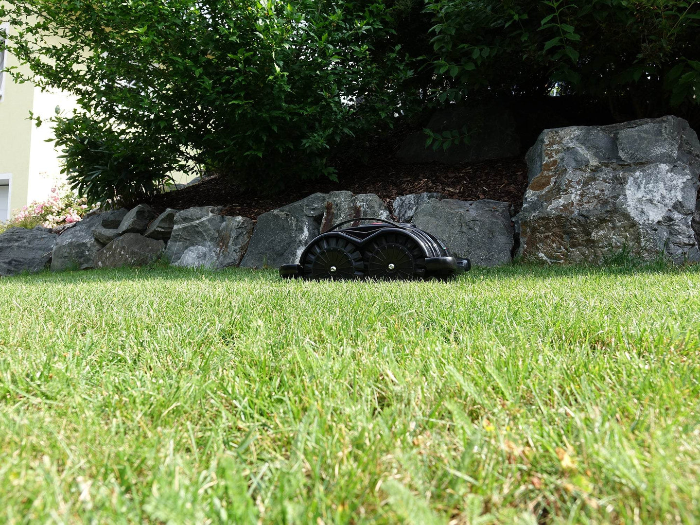 Das Bild zeigt den schwarzen Mähroboter Techline S6, der auf der Grasfläche steht im Hintergrund sind Büsche und eine Steindekoration.