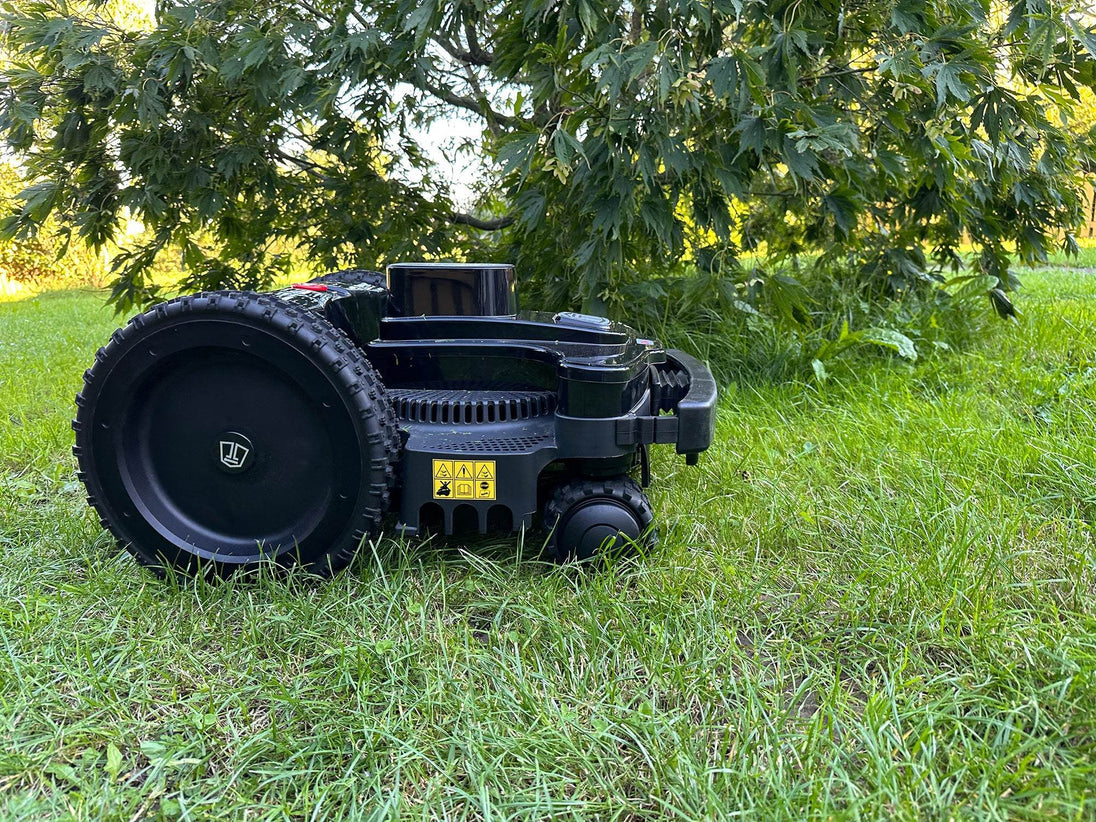Eine Nahaufnahme des Techline BX4 4WD Mähroboters im Gras, mit besonderem Fokus auf das große, schwarze Antriebsrad. Das robuste Profil des Rades ist deutlich erkennbar. Am Gehäuse sind gelbe Warnschilder mit Sicherheitshinweisen angebracht. Der Roboter steht auf saftig grünem Rasen, und im Hintergrund sind die Blätter eines Baumes zu sehen, die vom warmen Abendlicht durchflutet werden. Die schwarze, hochwertige Konstruktion des Mähroboters kommt in dieser Detailaufnahme besonders gut zur Geltung.