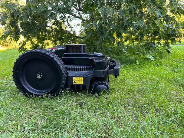 Eine Nahaufnahme des Techline BX4 4WD Mähroboters im Gras, mit besonderem Fokus auf das große, schwarze Antriebsrad. Das robuste Profil des Rades ist deutlich erkennbar. Am Gehäuse sind gelbe Warnschilder mit Sicherheitshinweisen angebracht. Der Roboter steht auf saftig grünem Rasen, und im Hintergrund sind die Blätter eines Baumes zu sehen, die vom warmen Abendlicht durchflutet werden. Die schwarze, hochwertige Konstruktion des Mähroboters kommt in dieser Detailaufnahme besonders gut zur Geltung.