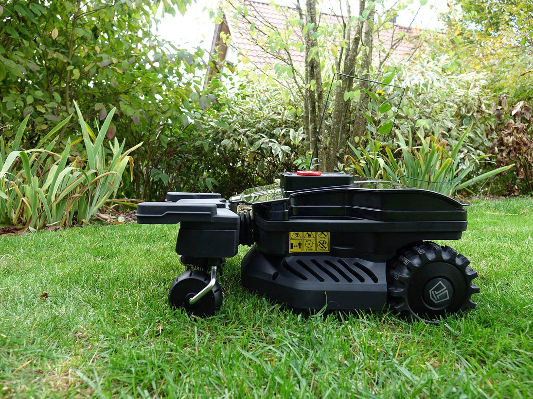 Das Bild zeigt den Mähroboter seitlich stehend vor einer Pflanze, wobei die Sicherheitskennzeichen sowie das Logo auf den Reifen deutlich zu erkennen sind.