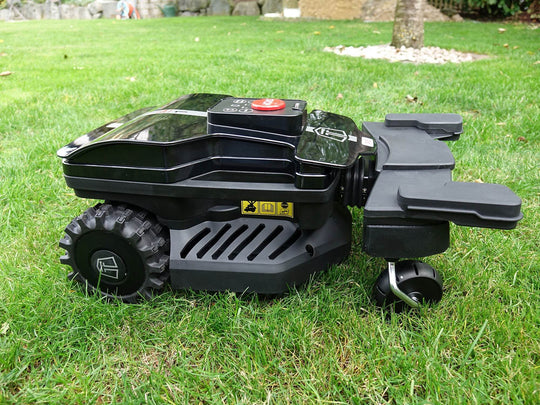 Das Bild zeigt den Mähroboter von der Seite, wobei das Kennzeichen sowie die breiten Räder und die Stoßsensoren deutlich zu erkennen sind.