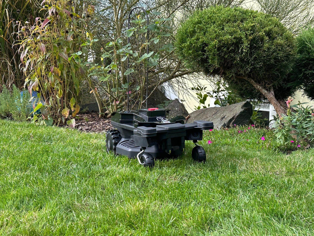 Das Bild zeigt einen schwarzen Techline NEXT TECH ZR EVO Mähroboter in der Frontansicht. Im Hintergrund sind Sträucher zu sehen.