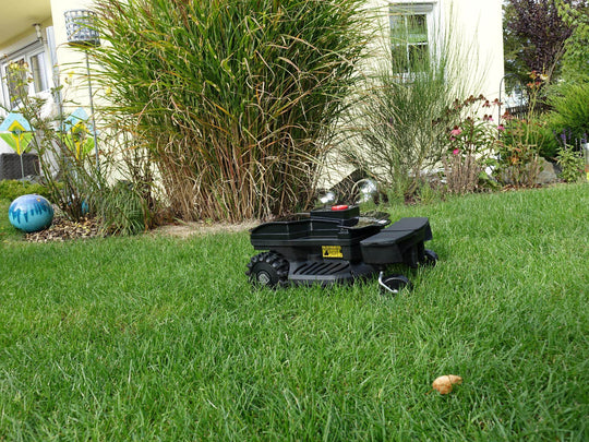 Das Bild zeigt den schwarzen Mähroboter Techline ZR in einer seitlichen Ansicht, wie er auf einer Gartenwiese steht. Im Hintergrund sind die Terrasse, Büsche, Blumen und Steine zu sehen.