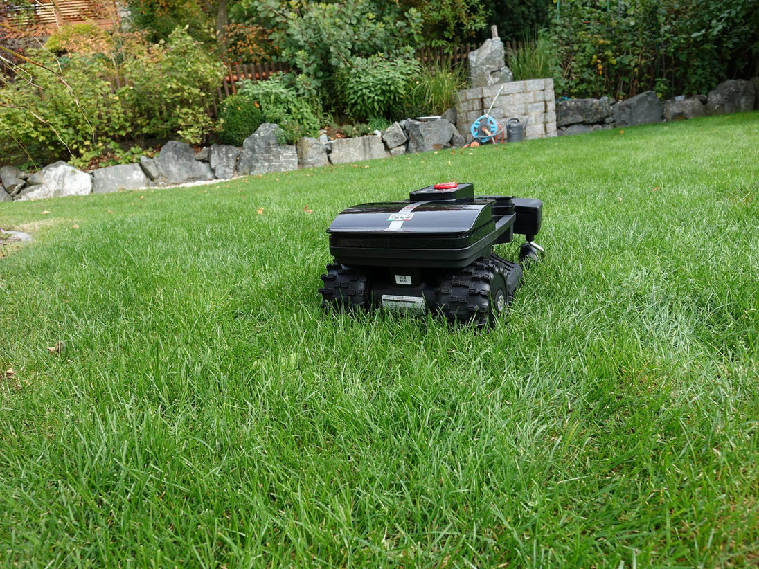 Das Bild zeigt den schwarzen Mähroboter Techline ZR in einer hinteren Ansicht, wie er auf einer Gartenwiese steht. Im Hintergrund sind ein Brunnen und ein Gartenschlauch zu sehen.
