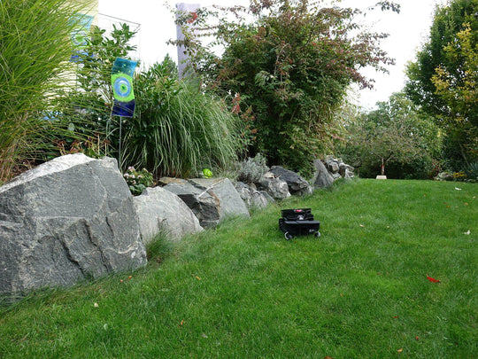 Das Bild zeigt den schwarzen Mähroboter Techline ZR aus der Vogelperspektive, wie er einen Abhang hinunterfährt. Hinter ihm sind Gartendeko-Elemente zu sehen, die eine gepflegte und ästhetische Gartenatmosphäre schaffen. Die Aufnahme unterstreicht die Geländetauglichkeit und Stabilität des Mähroboters, selbst bei unebenen Flächen wie einem Abhang.