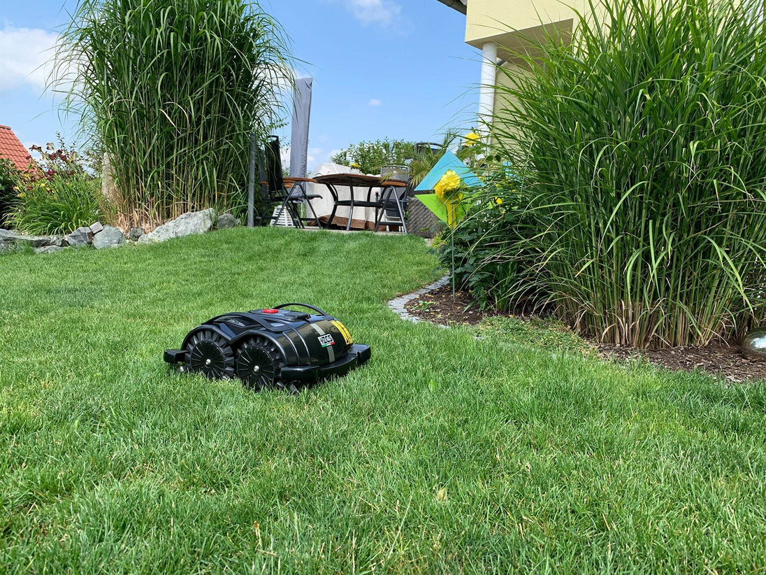 Das Bild zeigt den schwarzen Mähroboter Techline S6, der vor einer Terrasse steht und den Rasen mäht. Der Roboter ist in Aktion zu sehen, während er präzise und effizient arbeitet. Die Terrasse im Hintergrund verleiht der Szene einen häuslichen und praktischen Charakter, der die Einsatzmöglichkeiten des Geräts im Garten unterstreicht.