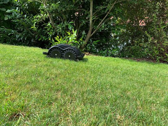 Das Bild zeigt den schwarzen Mähroboter Techline S6, der einen kleinen Abhang hinunterfährt. Im Hintergrund sind Bäume und Gebüsch zu sehen. Die Aufnahme unterstreicht die Geländetauglichkeit und Stabilität des Mähroboters, selbst bei unebenen Flächen.