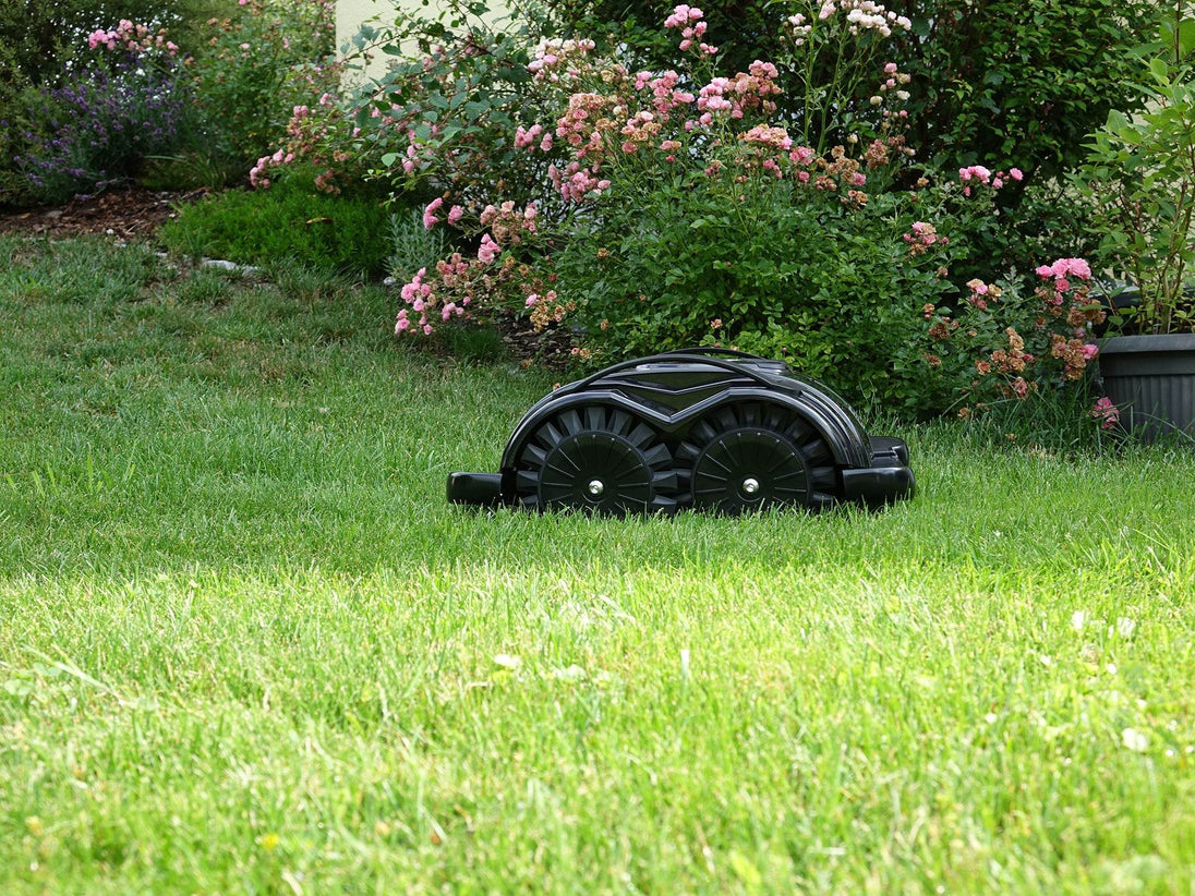 Das Bild zeigt den schwarzen Mähroboter Techline S6, der auf einer Wiese steht und von üppigen Büschen sowie einem Rosenstrauch umgeben ist.