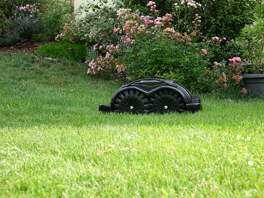Das Bild zeigt den schwarzen Mähroboter Techline S6, der auf einer Wiese steht und von üppigen Büschen sowie einem Rosenstrauch umgeben ist.